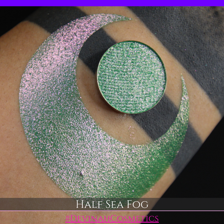 A round eyeshadow pan labeled 'HALF SEA FOG' made by Devinah Cosmetics, placed over a large crescent moon swatch of the same shimmering shifty eyeshadow on skin. The eyeshadow has a duochrome multichrome shift between colors lime green, yellow, pink.