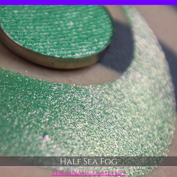 A round eyeshadow pan labeled 'HALF SEA FOG' made by Devinah Cosmetics, placed over a large crescent moon swatch of the same shimmering shifty eyeshadow on skin. The eyeshadow has a duochrome multichrome shift between colors lime green, yellow, pink.