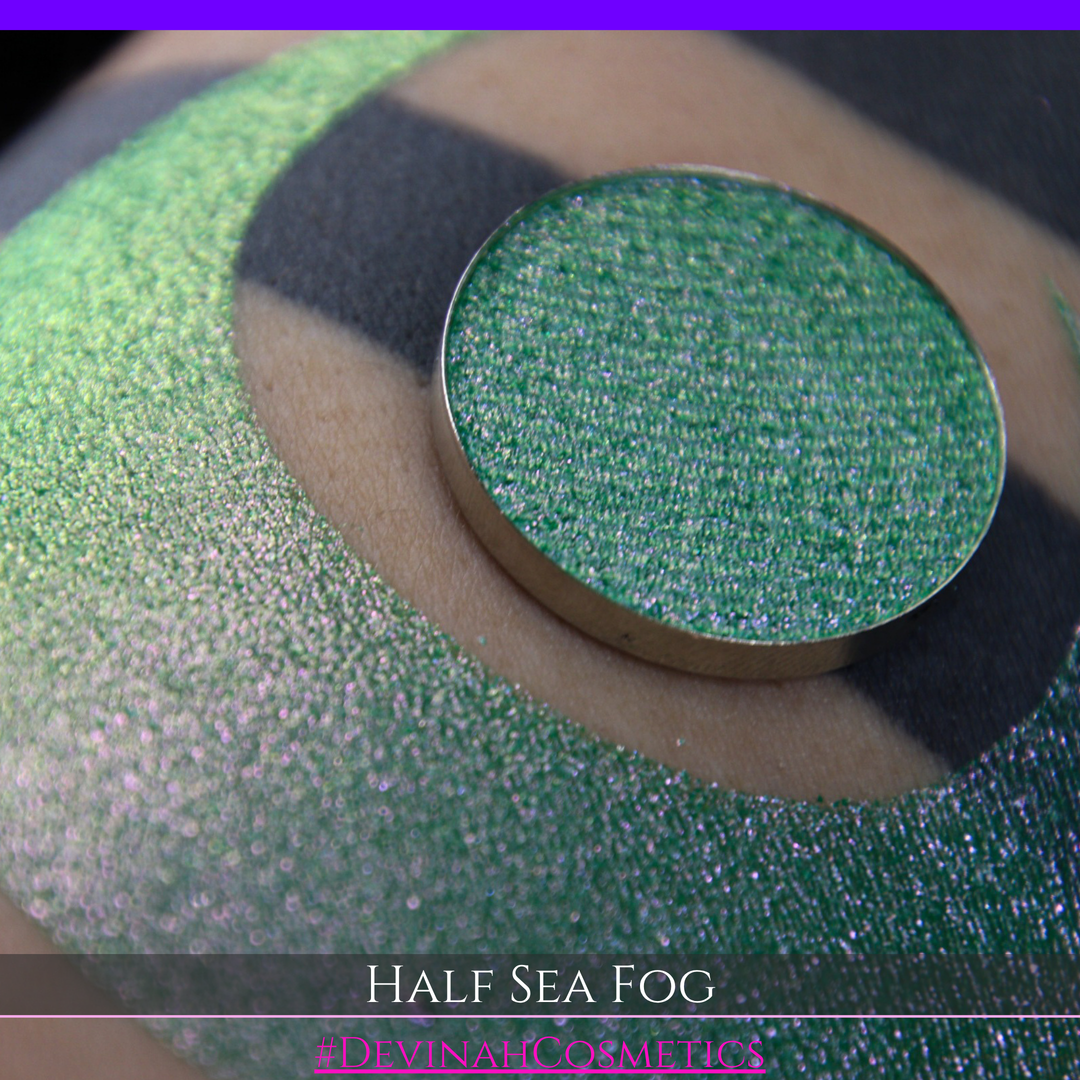 A round eyeshadow pan labeled 'HALF SEA FOG' made by Devinah Cosmetics, placed over a large crescent moon swatch of the same shimmering shifty eyeshadow on skin. The eyeshadow has a duochrome multichrome shift between colors lime green, yellow, pink.