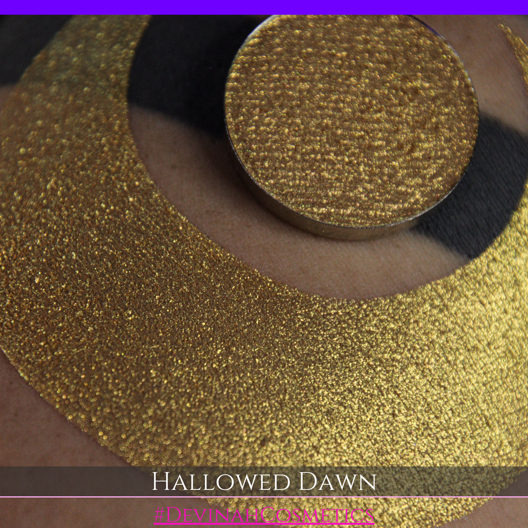 A close-up of a person's arm with a large gold crescent moon eyeshadow makeup swatch and a matching round gold eyeshadow pan, with text reading 'HALLOWED DAWN' and '#DEVINAHCOSMETICS' at the bottom.