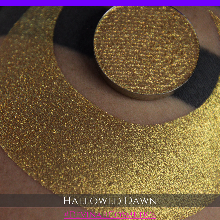 A close-up of a person's arm with a large gold crescent moon eyeshadow makeup swatch and a matching round gold eyeshadow pan, with text reading 'HALLOWED DAWN' and '#DEVINAHCOSMETICS' at the bottom.