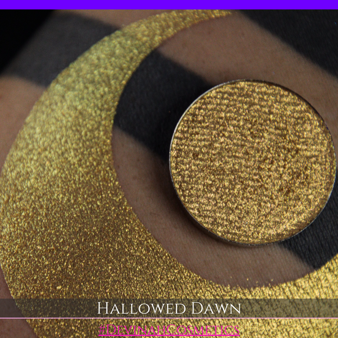 A close-up of a person's arm with a large gold crescent moon eyeshadow makeup swatch and a matching round gold eyeshadow pan, with text reading 'HALLOWED DAWN' and '#DEVINAHCOSMETICS' at the bottom.