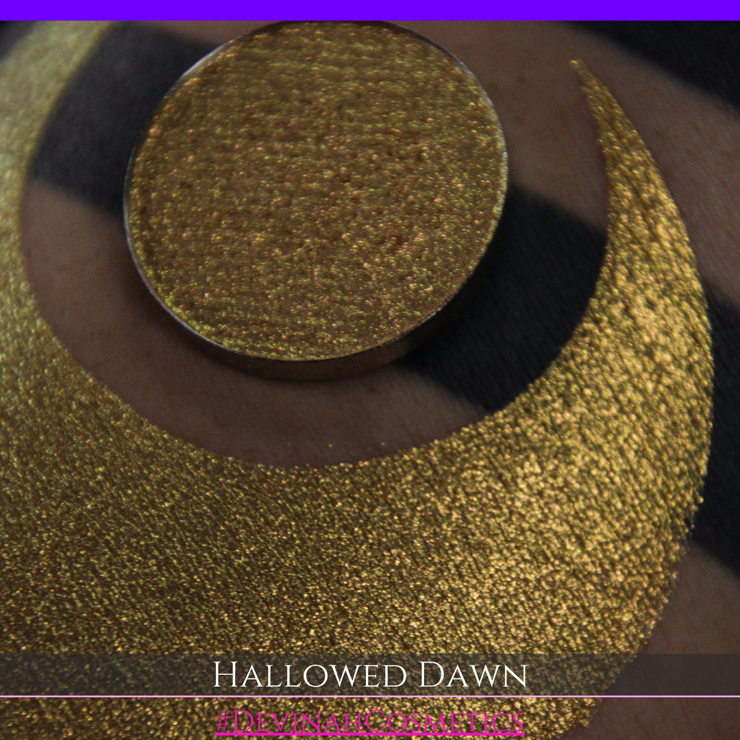 A close-up of a person's arm with a large gold crescent moon eyeshadow makeup swatch and a matching round gold eyeshadow pan, with text reading 'HALLOWED DAWN' and '#DEVINAHCOSMETICS' at the bottom.