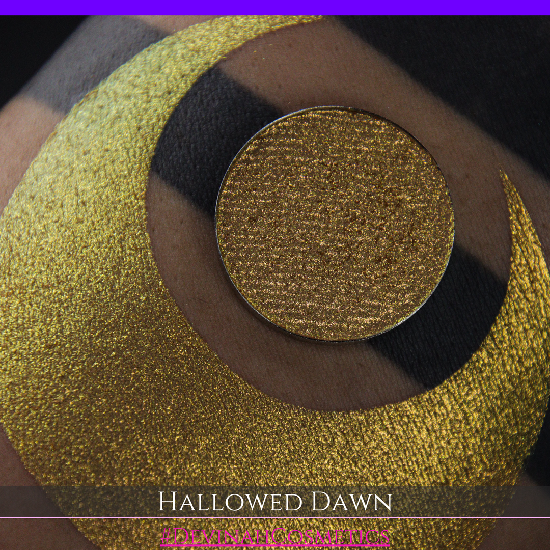 A close-up of a person's arm with a large gold crescent moon eyeshadow makeup swatch and a matching round gold eyeshadow pan, with text reading 'HALLOWED DAWN' and '#DEVINAHCOSMETICS' at the bottom.