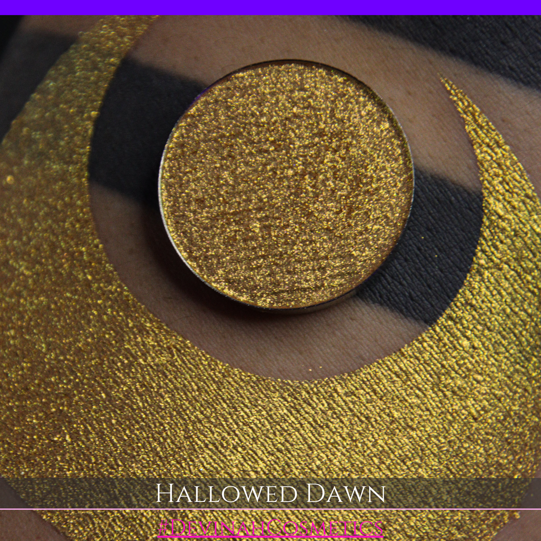 A close-up of a person's arm with a large gold crescent moon eyeshadow makeup swatch and a matching round gold eyeshadow pan, with text reading 'HALLOWED DAWN' and '#DEVINAHCOSMETICS' at the bottom.