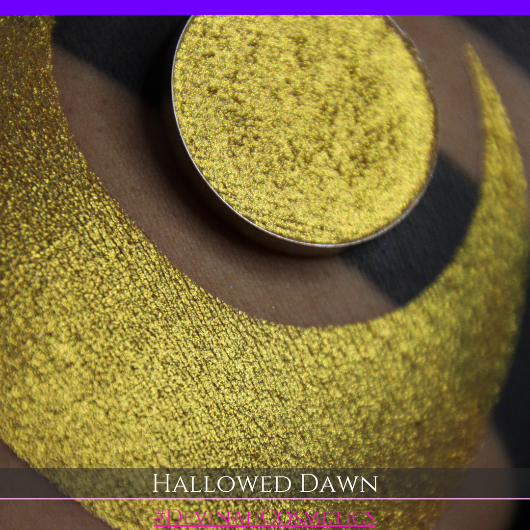 A close-up of a person's arm with a large gold crescent moon eyeshadow makeup swatch and a matching round gold eyeshadow pan, with text reading 'HALLOWED DAWN' and '#DEVINAHCOSMETICS' at the bottom.