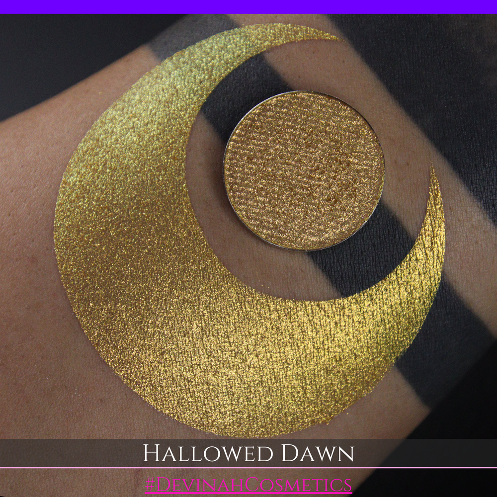 A close-up of a person's arm with a large gold crescent moon eyeshadow makeup swatch and a matching round gold eyeshadow pan, with text reading 'HALLOWED DAWN' and '#DEVINAHCOSMETICS' at the bottom.