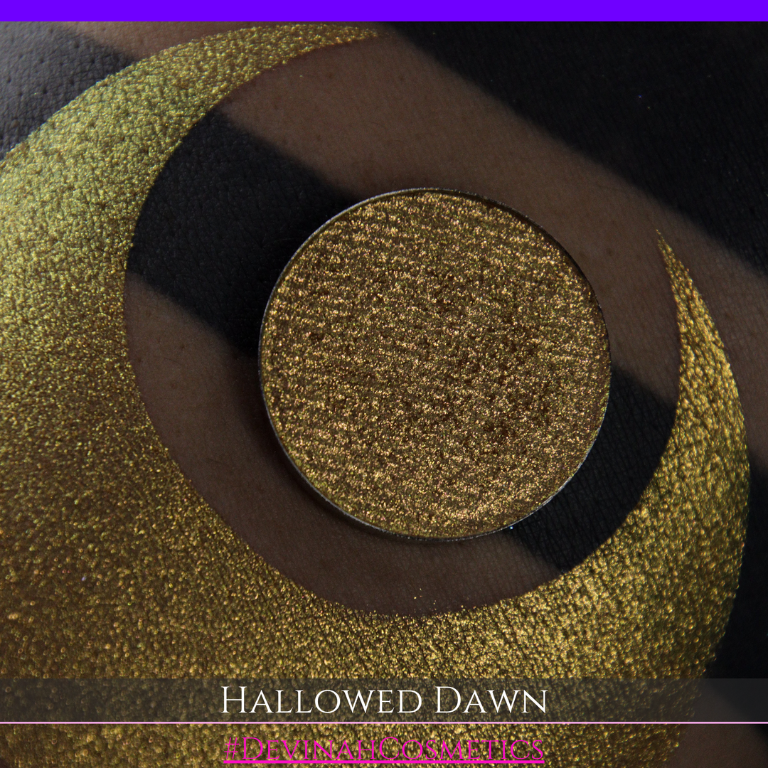 A close-up of a person's arm with a large gold crescent moon eyeshadow makeup swatch and a matching round gold eyeshadow pan, with text reading 'HALLOWED DAWN' and '#DEVINAHCOSMETICS' at the bottom.