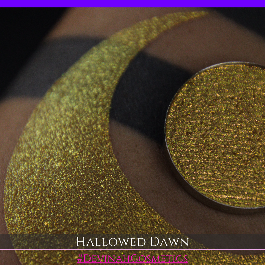 A close-up of a person's arm with a large gold crescent moon eyeshadow makeup swatch and a matching round gold eyeshadow pan, with text reading 'HALLOWED DAWN' and '#DEVINAHCOSMETICS' at the bottom.