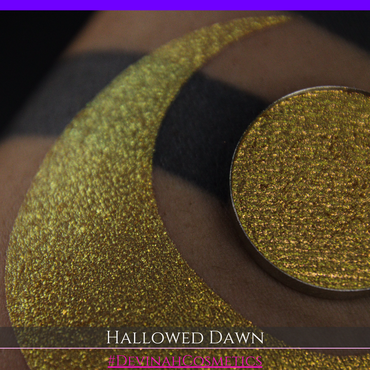 A close-up of a person's arm with a large gold crescent moon eyeshadow makeup swatch and a matching round gold eyeshadow pan, with text reading 'HALLOWED DAWN' and '#DEVINAHCOSMETICS' at the bottom.