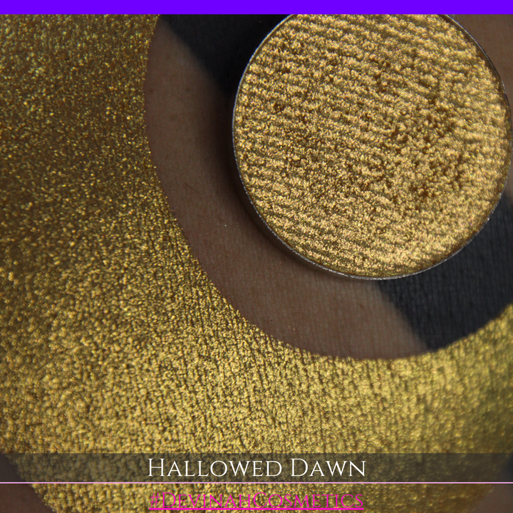 A close-up of a person's arm with a large gold crescent moon eyeshadow makeup swatch and a matching round gold eyeshadow pan, with text reading 'HALLOWED DAWN' and '#DEVINAHCOSMETICS' at the bottom.