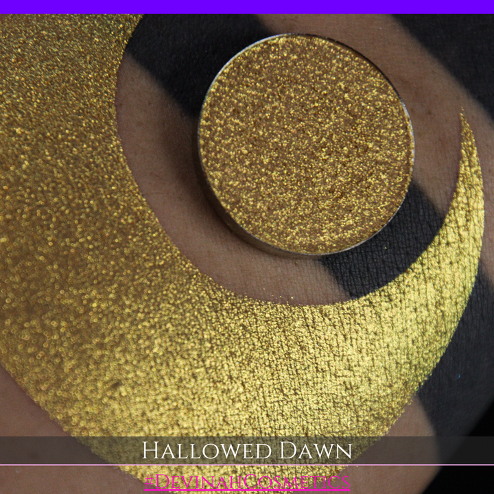 A close-up of a person's arm with a large gold crescent moon eyeshadow makeup swatch and a matching round gold eyeshadow pan, with text reading 'HALLOWED DAWN' and '#DEVINAHCOSMETICS' at the bottom.