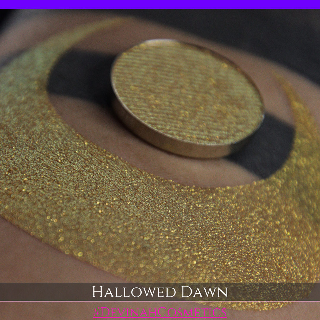 A close-up of a person's arm with a large gold crescent moon eyeshadow makeup swatch and a matching round gold eyeshadow pan, with text reading 'HALLOWED DAWN' and '#DEVINAHCOSMETICS' at the bottom.