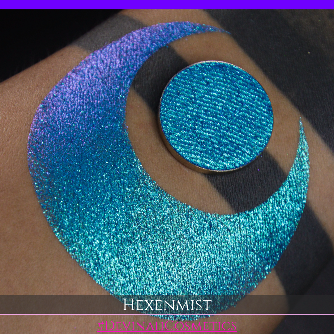 A close-up of a large crescent swatch of shimmering eyeshadow makeup in bright teal, aqua, and purple tones. The text 'HEXENMIST' and '#DEVINAHCOSMETICS' appear at the bottom.