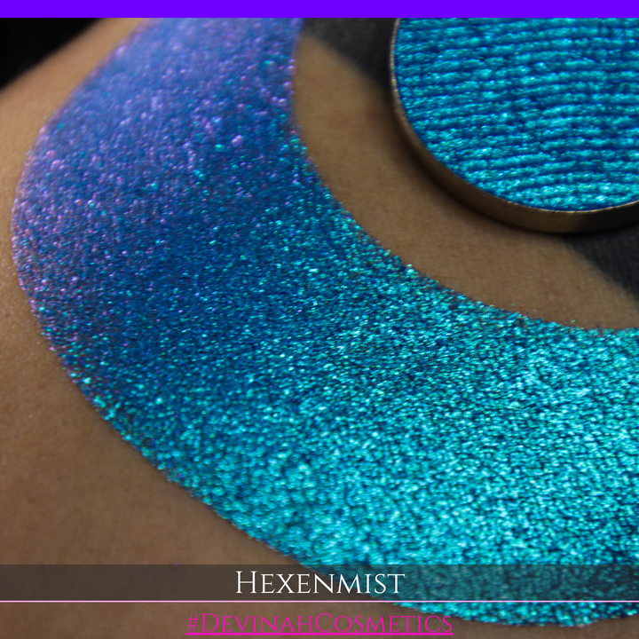 A close-up of a large crescent swatch of shimmering eyeshadow makeup in bright teal, aqua, and purple tones. The text 'HEXENMIST' and '#DEVINAHCOSMETICS' appear at the bottom.