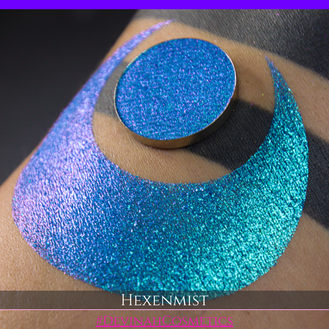 A close-up of a large crescent swatch of shimmering eyeshadow makeup in bright teal, aqua, and purple tones. The text 'HEXENMIST' and '#DEVINAHCOSMETICS' appear at the bottom.