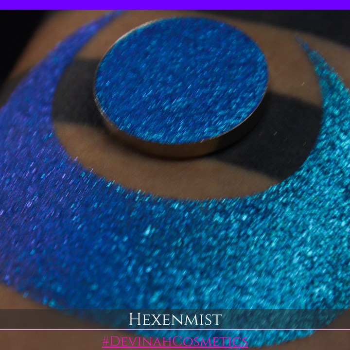A close-up of a large crescent swatch of shimmering eyeshadow makeup in bright teal, aqua, and purple tones. The text 'HEXENMIST' and '#DEVINAHCOSMETICS' appear at the bottom.
