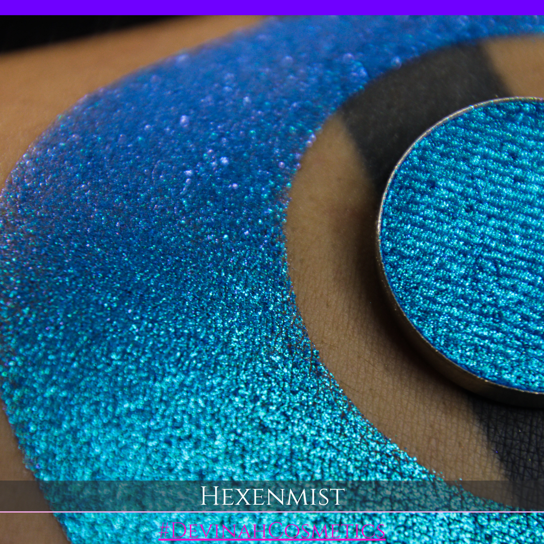 A close-up of a large crescent swatch of shimmering eyeshadow makeup in bright teal, aqua, and purple tones. The text 'HEXENMIST' and '#DEVINAHCOSMETICS' appear at the bottom.