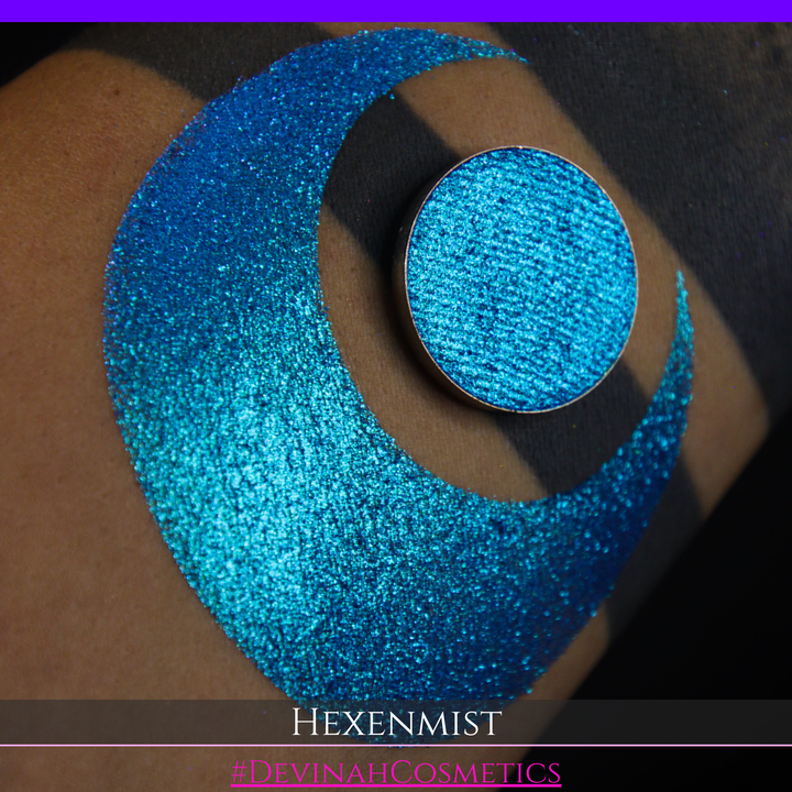 A close-up of a large crescent swatch of shimmering eyeshadow makeup in bright teal, aqua, and purple tones. The text 'HEXENMIST' and '#DEVINAHCOSMETICS' appear at the bottom.