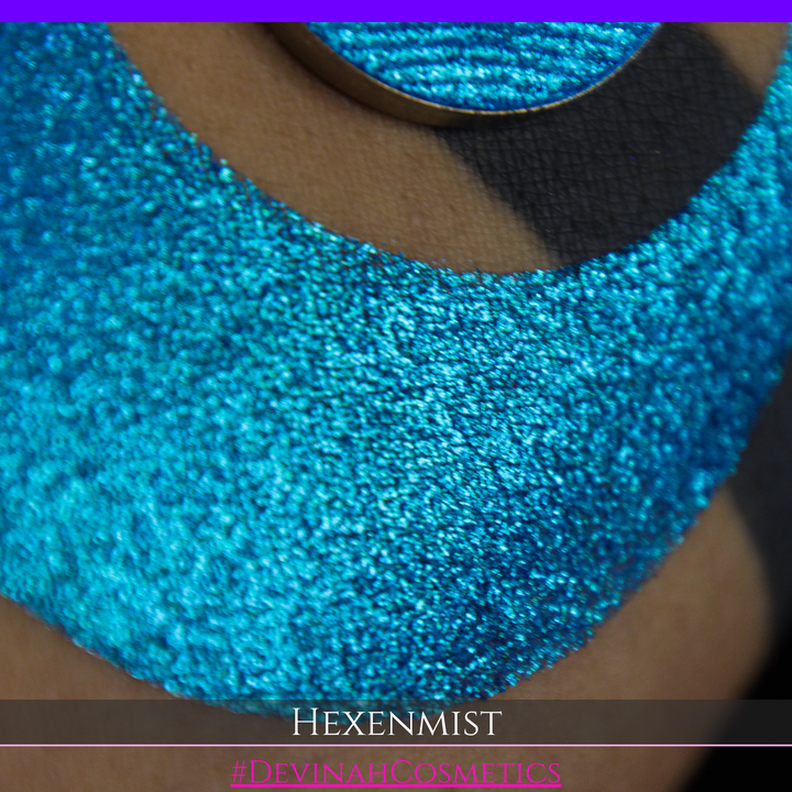 A close-up of a large crescent swatch of shimmering eyeshadow makeup in bright teal, aqua, and purple tones. The text 'HEXENMIST' and '#DEVINAHCOSMETICS' appear at the bottom.