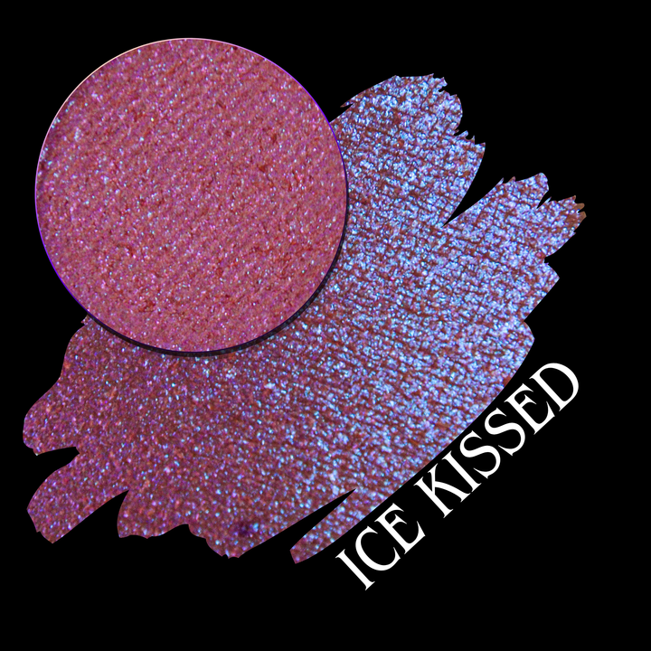 A circular makeup eyeshadow pan of shimmering pinkish-bluish sparkle is shown over a swatch on a black background, with the text 'ICE KISSED' in white at the lower right made by Devinah Cosmetics.