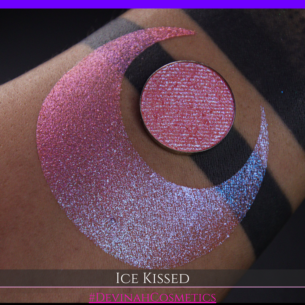 A close-up of a person's arm with a large crescent moon eyeshadow swatch and circular makeup eyeshadow pan of shimmery pink, orange and blue sparkle, labeled 'Ice Kissed' by Devinah Cosmetics.