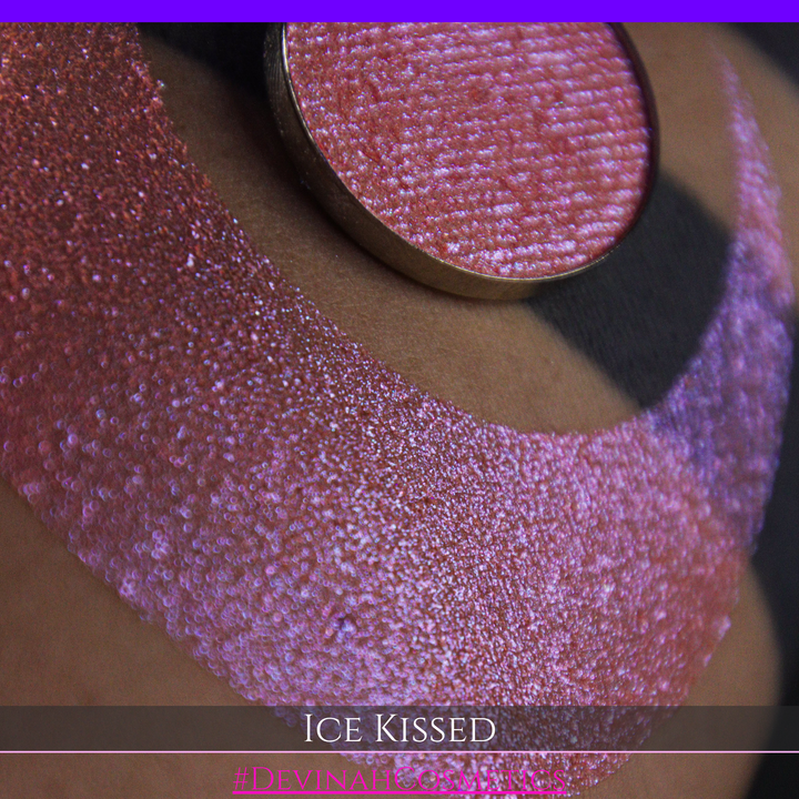 A close-up of a person's arm with a large crescent moon eyeshadow swatch and circular makeup eyeshadow pan of shimmery pink, orange and blue sparkle, labeled 'Ice Kissed' by Devinah Cosmetics.