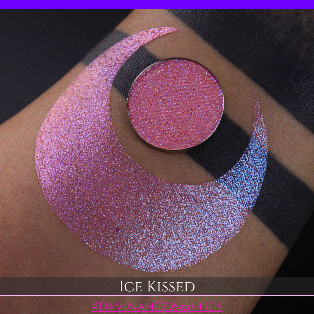 A close-up of a person's arm with a large crescent moon eyeshadow swatch and circular makeup eyeshadow pan of shimmery pink, orange and blue sparkle, labeled 'Ice Kissed' by Devinah Cosmetics.
