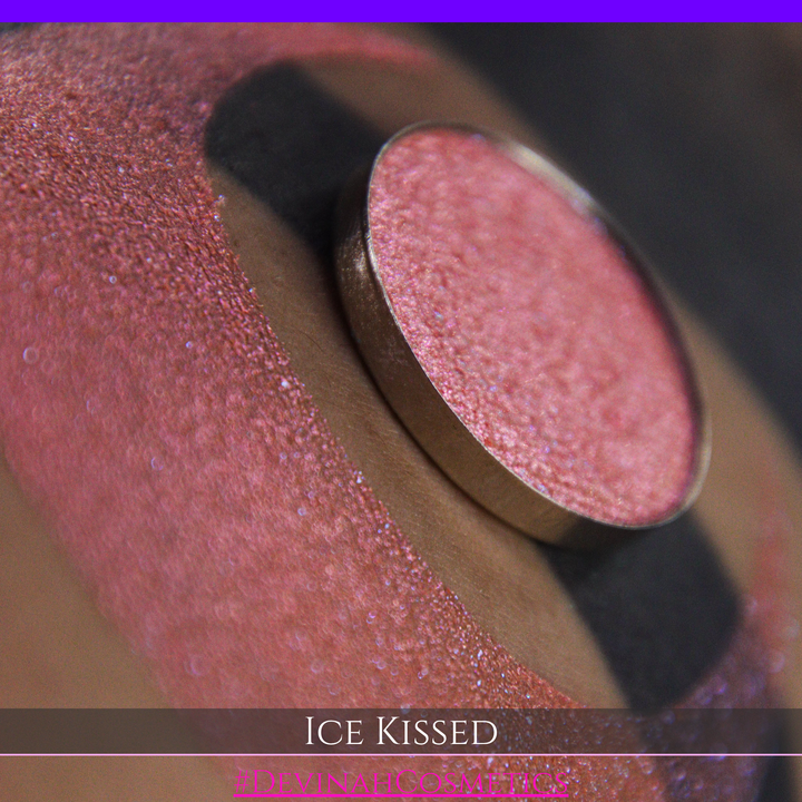 A close-up of a person's arm with a large crescent moon eyeshadow swatch and circular makeup eyeshadow pan of shimmery pink, orange and blue sparkle, labeled 'Ice Kissed' by Devinah Cosmetics.