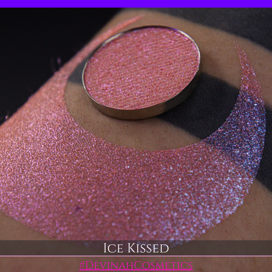 A close-up of a person's arm with a large crescent moon eyeshadow swatch and circular makeup eyeshadow pan of shimmery pink, orange and blue sparkle, labeled 'Ice Kissed' by Devinah Cosmetics.