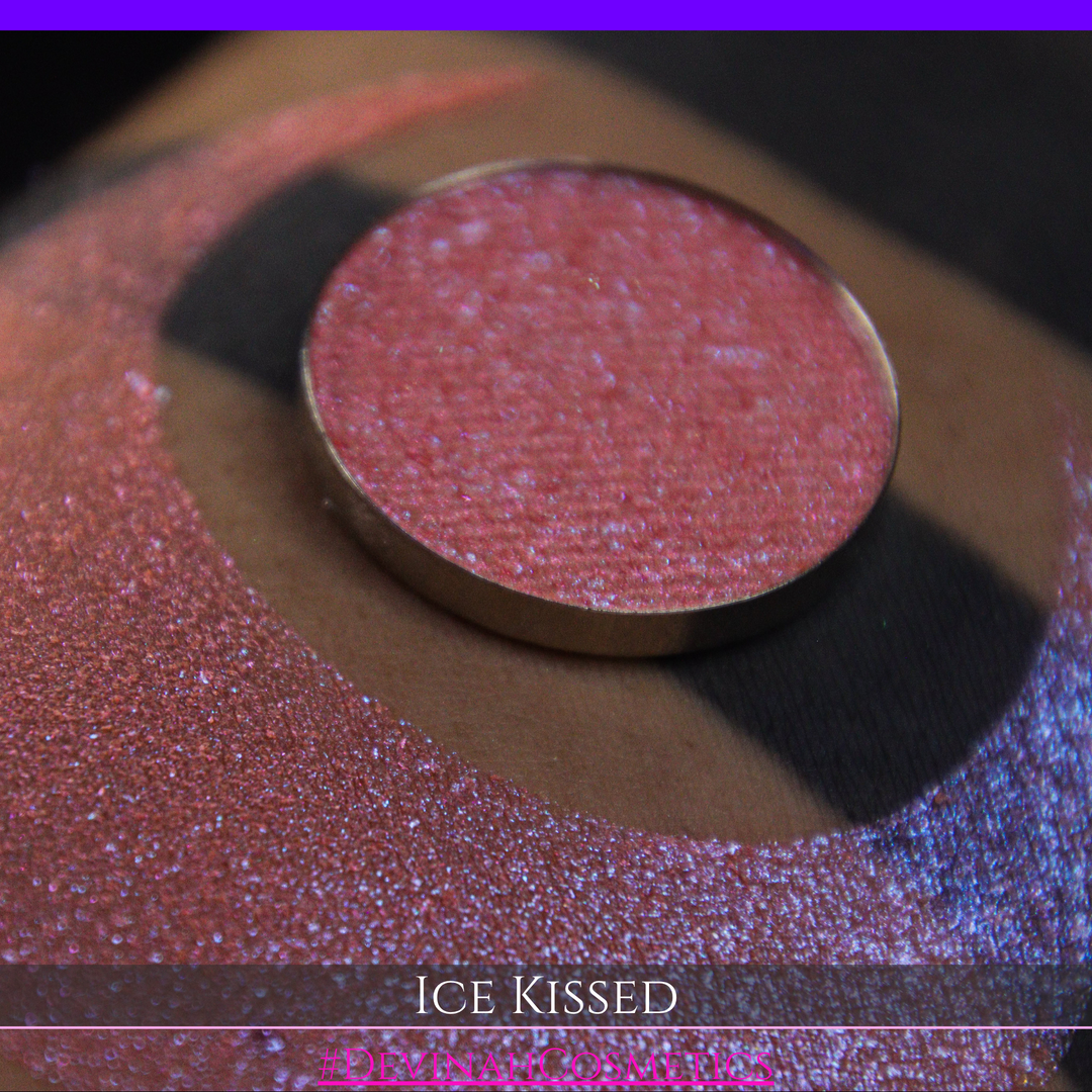 A close-up of a person's arm with a large crescent moon eyeshadow swatch and circular makeup eyeshadow pan of shimmery pink, orange and blue sparkle, labeled 'Ice Kissed' by Devinah Cosmetics.