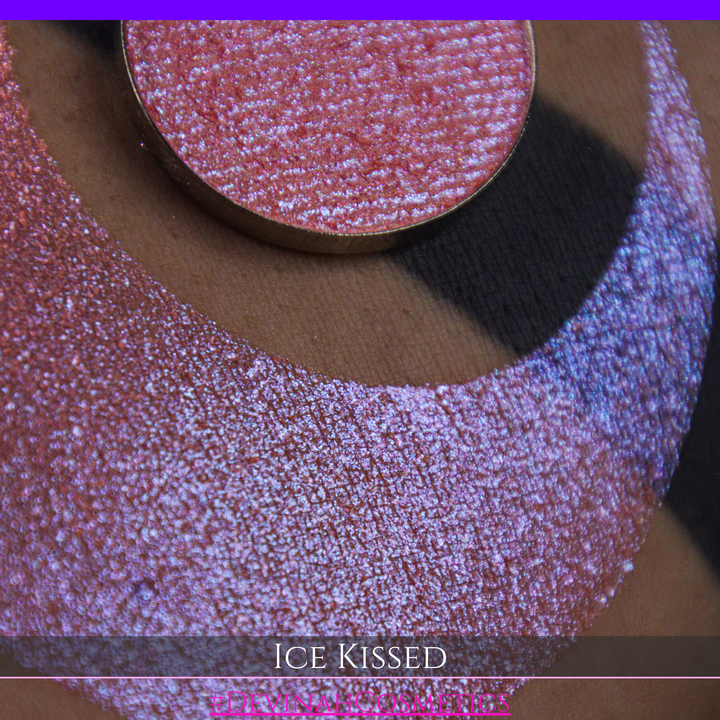 A close-up of a person's arm with a large crescent moon eyeshadow swatch and circular makeup eyeshadow pan of shimmery pink, orange and blue sparkle, labeled 'Ice Kissed' by Devinah Cosmetics.