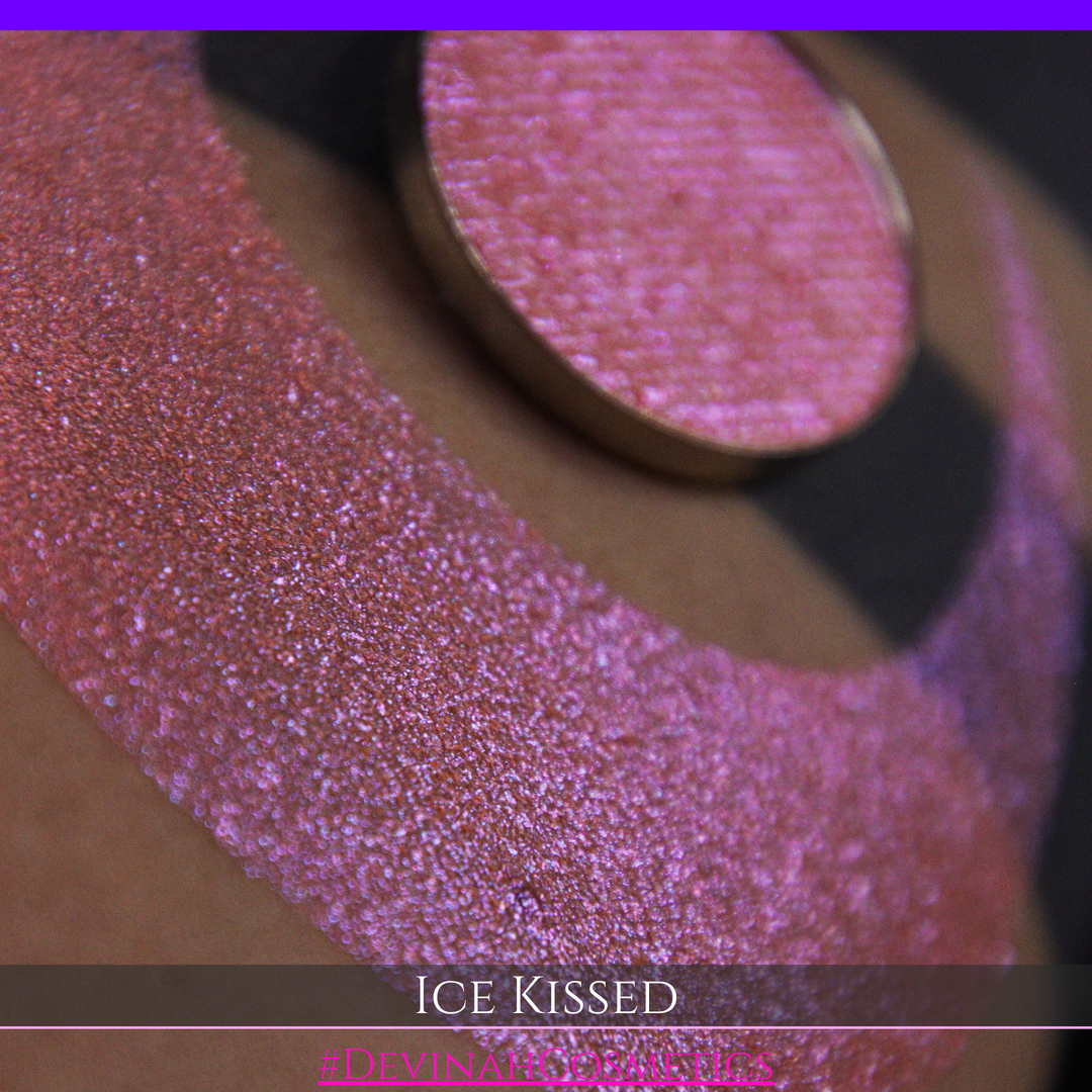 A close-up of a person's arm with a large crescent moon eyeshadow swatch and circular makeup eyeshadow pan of shimmery pink, orange and blue sparkle, labeled 'Ice Kissed' by Devinah Cosmetics.