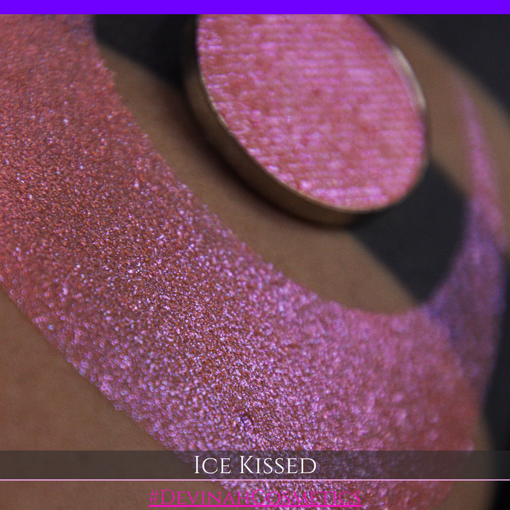 A close-up of a person's arm with a large crescent moon eyeshadow swatch and circular makeup eyeshadow pan of shimmery pink, orange and blue sparkle, labeled 'Ice Kissed' by Devinah Cosmetics.