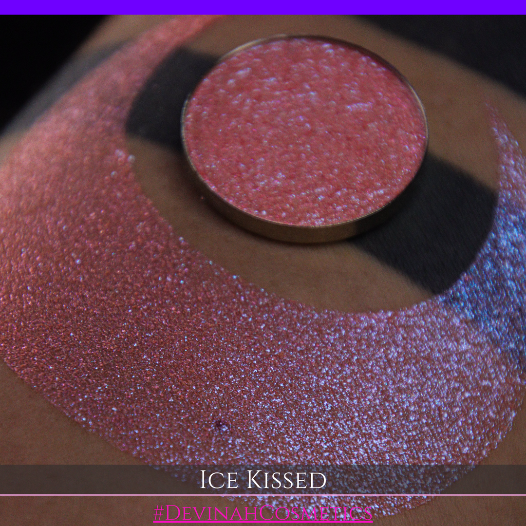 A close-up of a person's arm with a large crescent moon eyeshadow swatch and circular makeup eyeshadow pan of shimmery pink, orange and blue sparkle, labeled 'Ice Kissed' by Devinah Cosmetics.