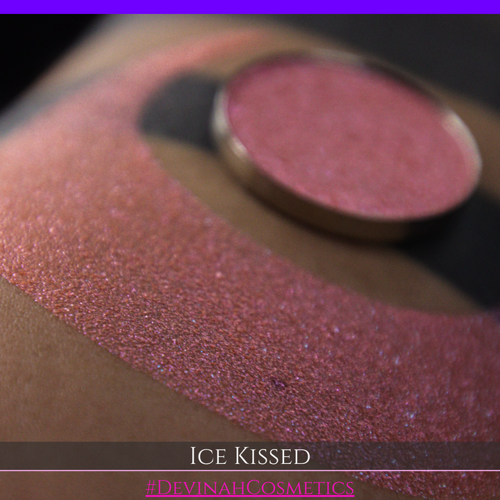 A close-up of a person's arm with a large crescent moon eyeshadow swatch and circular makeup eyeshadow pan of shimmery pink, orange and blue sparkle, labeled 'Ice Kissed' by Devinah Cosmetics.