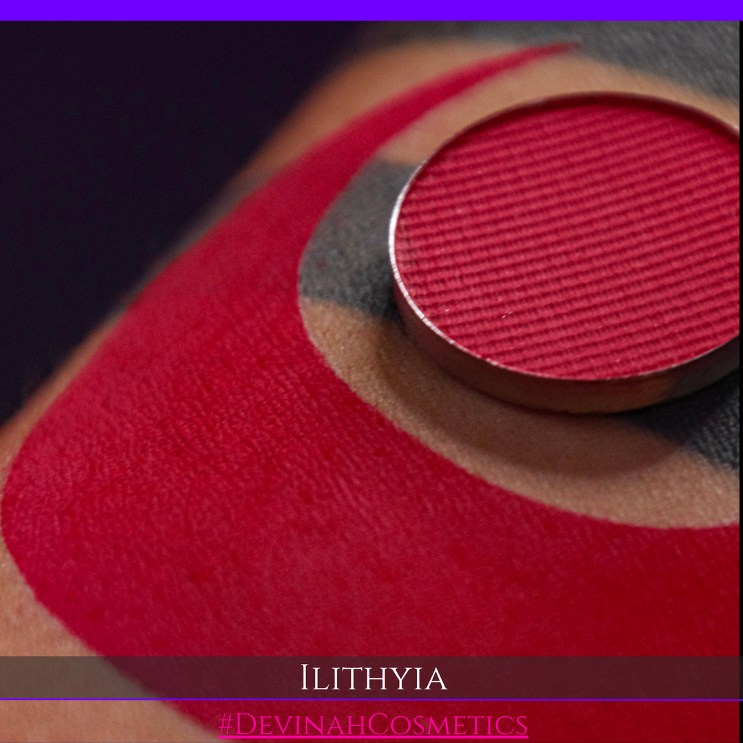 A close-up of a makeup product pan in a deep berry red shade resting on top of a red eyeshadow swatch on skin, with the brand name ILITHYIA and hashtag #DEVINAHCOSMETICS visible at the bottom.