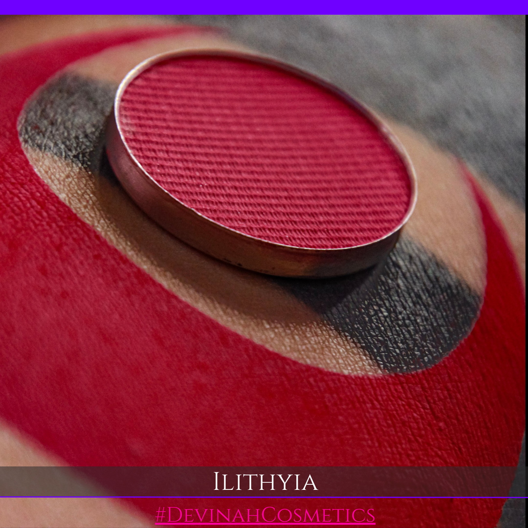 A close-up of a round makeup pan containing vibrant red powder, swatched on tan skin. The name 'ILITHYIA' and '#DEVINAHCOSMETICS' are displayed at the bottom.