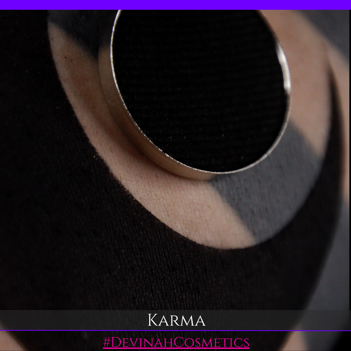 A close-up of a black circular cosmetic pan or compact with a silver-toned rim, resting on a black textured surface. A beige circular shape is visible behind it, and the text 'KARMA' and '#DEVINAHCOSMETICS' appears at the bottom.