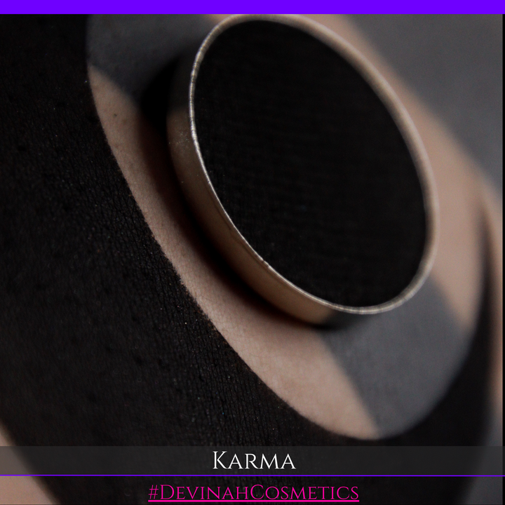 A close-up of a black circular cosmetic pan or compact with a metallic rim, resting on a beige and black textured surface. Text at the bottom reads 'KARMA' and '#DEVINAHCOSMETICS'.
