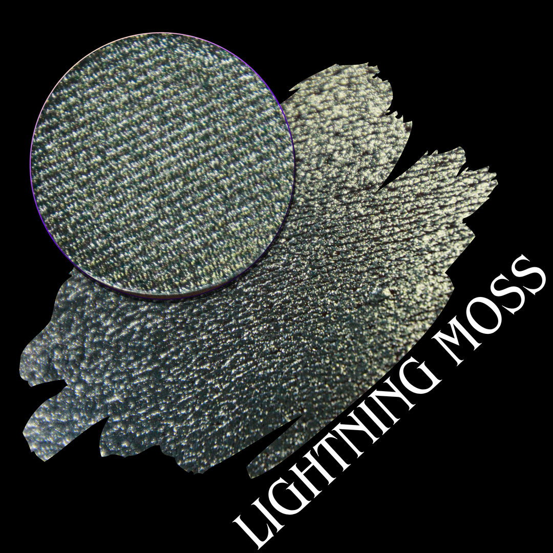 A round eyeshadow pan labeled 'LIGHTNING MOSS' made by Devinah Cosmetics, placed over a large crescent moon swatch of the same shimmering shifty eyeshadow on skin. The eyeshadow has a duochrome multichrome shift between colors pewter, silver, blue, green.