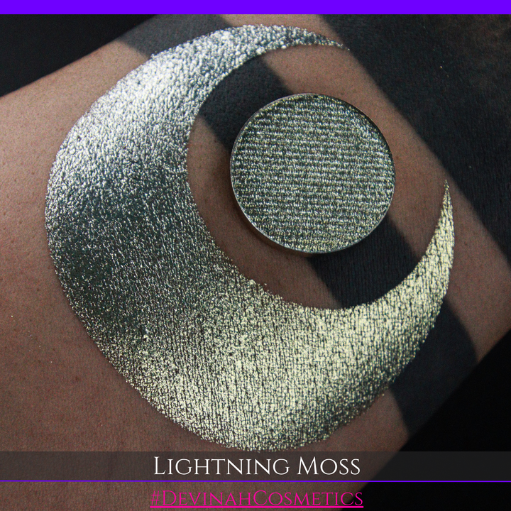A round eyeshadow pan labeled 'LIGHTNING MOSS' made by Devinah Cosmetics, placed over a large crescent moon swatch of the same shimmering shifty eyeshadow on skin. The eyeshadow has a duochrome multichrome shift between colors pewter, silver, blue, green.