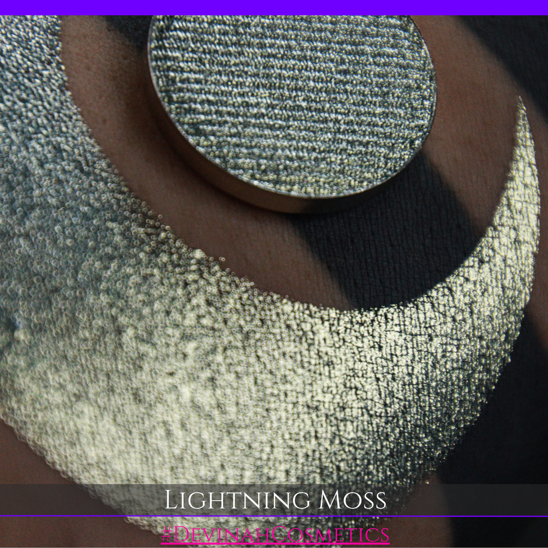 A round eyeshadow pan labeled 'LIGHTNING MOSS' made by Devinah Cosmetics, placed over a large crescent moon swatch of the same shimmering shifty eyeshadow on skin. The eyeshadow has a duochrome multichrome shift between colors pewter, silver, blue, green.