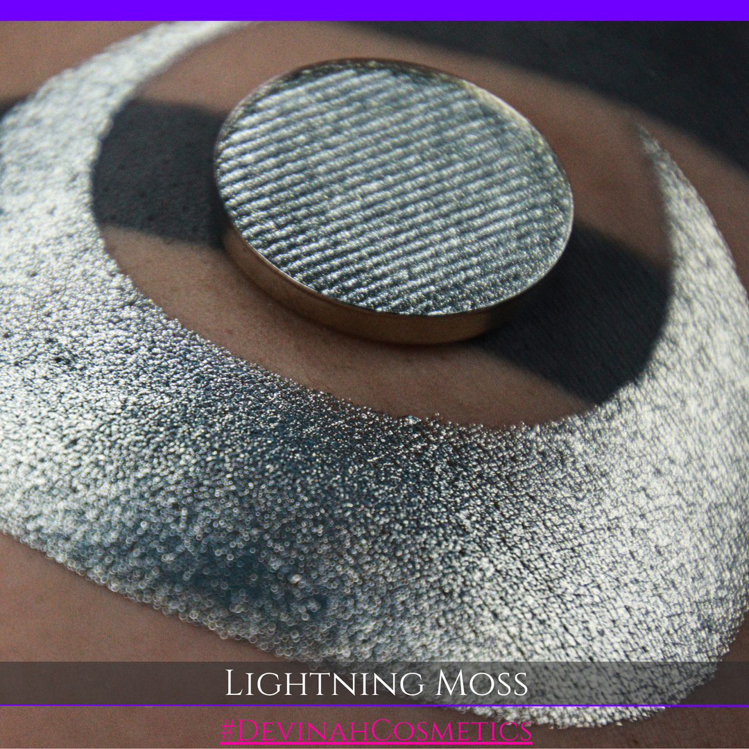 A round eyeshadow pan labeled 'LIGHTNING MOSS' made by Devinah Cosmetics, placed over a large crescent moon swatch of the same shimmering shifty eyeshadow on skin. The eyeshadow has a duochrome multichrome shift between colors pewter, silver, blue, green.