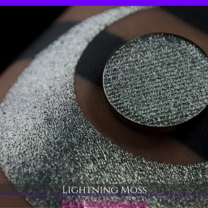 A round eyeshadow pan labeled 'LIGHTNING MOSS' made by Devinah Cosmetics, placed over a large crescent moon swatch of the same shimmering shifty eyeshadow on skin. The eyeshadow has a duochrome multichrome shift between colors pewter, silver, blue, green.