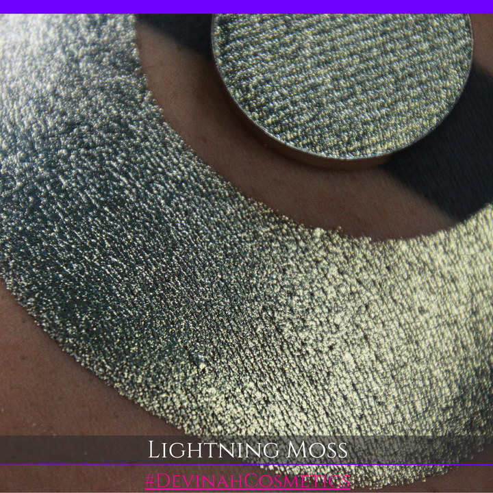 A round eyeshadow pan labeled 'LIGHTNING MOSS' made by Devinah Cosmetics, placed over a large crescent moon swatch of the same shimmering shifty eyeshadow on skin. The eyeshadow has a duochrome multichrome shift between colors pewter, silver, blue, green.
