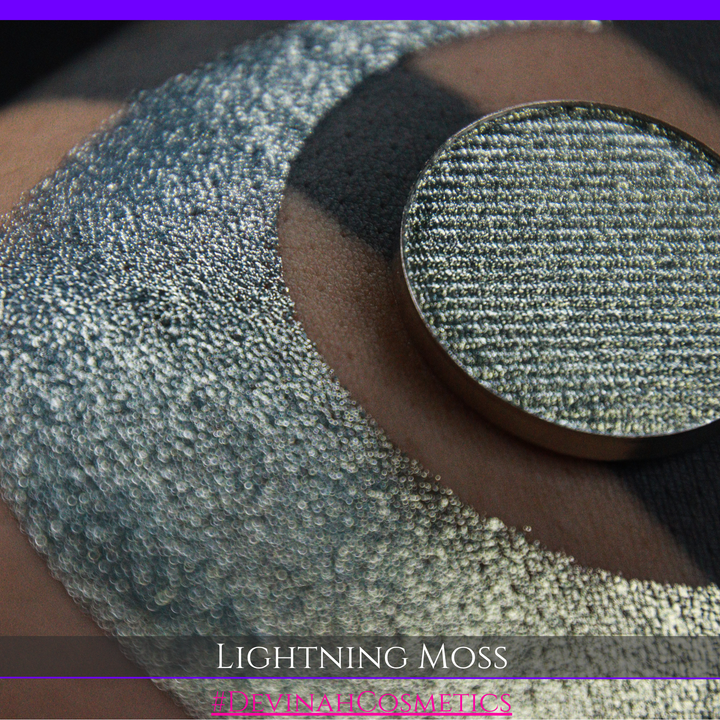 A round eyeshadow pan labeled 'LIGHTNING MOSS' made by Devinah Cosmetics, placed over a large crescent moon swatch of the same shimmering shifty eyeshadow on skin. The eyeshadow has a duochrome multichrome shift between colors pewter, silver, blue, green.