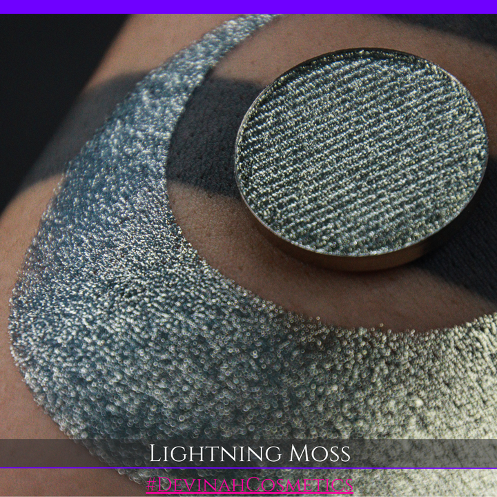A round eyeshadow pan labeled 'LIGHTNING MOSS' made by Devinah Cosmetics, placed over a large crescent moon swatch of the same shimmering shifty eyeshadow on skin. The eyeshadow has a duochrome multichrome shift between colors pewter, silver, blue, green.