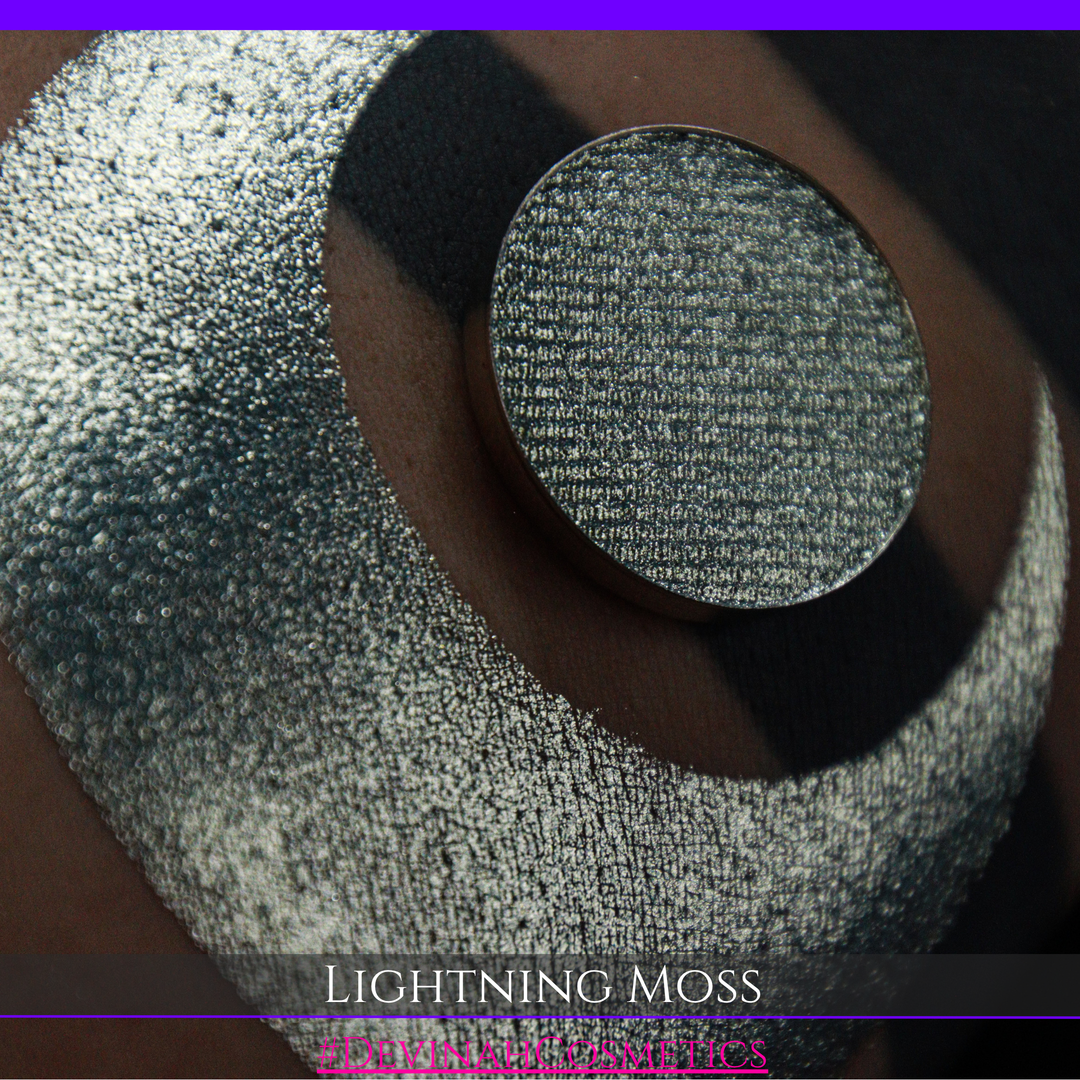 A round eyeshadow pan labeled 'LIGHTNING MOSS' made by Devinah Cosmetics, placed over a large crescent moon swatch of the same shimmering shifty eyeshadow on skin. The eyeshadow has a duochrome multichrome shift between colors pewter, silver, blue, green.