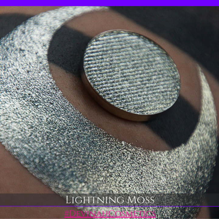 A round eyeshadow pan labeled 'LIGHTNING MOSS' made by Devinah Cosmetics, placed over a large crescent moon swatch of the same shimmering shifty eyeshadow on skin. The eyeshadow has a duochrome multichrome shift between colors pewter, silver, blue, green.